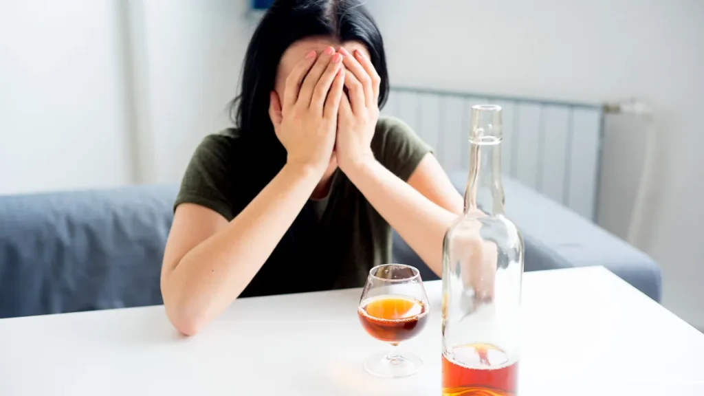 Woman covering her face at a table with a glass and bottle of alcohol