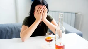 Woman covering her face at a table with a glass and bottle of alcohol
